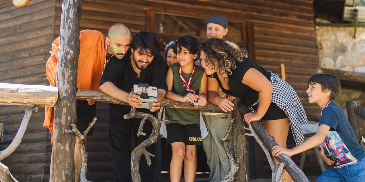 A diverse group of people joyfully watching drone footage on a wooden cabin porch.